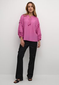 Blusa rosa con vestibilità ampia, dotata di maniche arricciate e colletto con fiocco e pon pon, abbinata a pantaloni neri a gamba dritta e sandali neri.