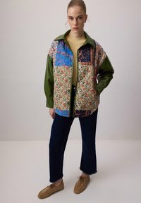 Quiltet patchwork jakke med grønne faux læderærmer, blomster- og geometriske mønstre i blå, beige og burgunderrød, med krave.