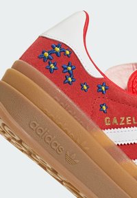 Baskets Adidas Gazelle rouges avec languette de talon blanche, semelle en gomme, bandes blanches et étoiles brodées bleues et jaunes sur le côté.