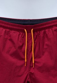Shorts de bain rouges avec une taille élastique, dotés d'une doublure intérieure bleu foncé et d'un cordon de serrage orange vif. Texture lisse sans motifs.