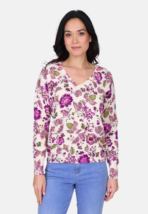 Femme portant un pull léger à col en V avec un motif floral violet et un jean bleu, debout devant un fond blanc uni.
