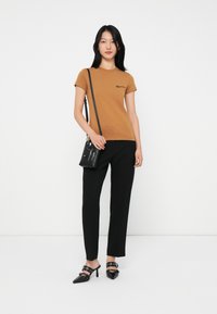 Bruine T-shirt met korte mouwen en een klein logo, gecombineerd met zwarte maatwerk broek en zwarte puntige hakken met banddetails. Inclusief schoudertas.
