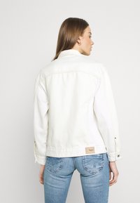 Vitt denimjacka med klassisk skärning, synlig sömnad och ett märkt lädermärke bak vid fållen. Bärs över blå jeans.