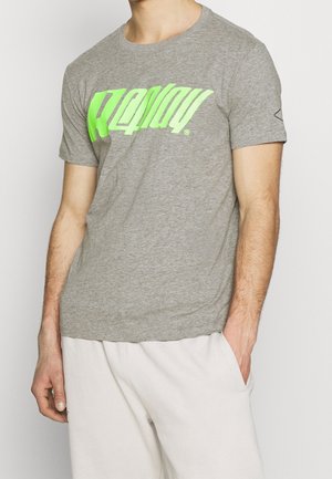 Homme portant un t-shirt gris avec un grand texte vert néon et un pantalon beige clair, debout avec une main dans la poche sur un fond blanc.