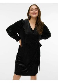 Robe portefeuille noire avec motif à pois brillants, décolleté en V, manches longues bouffantes, ceinture nouée à la taille et deux poches latérales. Texture lisse.