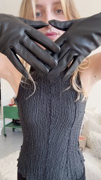 Melni ādas cimdi ar pieguļošu dizainu, gludu tekstūru un garām pirkstiem, novietoti uz teksturēta melna bezpiedurkņu topa.