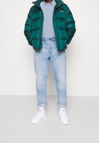 Zelená péřová bunda s lesklým povrchem, světle modrá košile na knoflíky, světle modré denimové džíny a bílé tenisky. Pohodlný outfit s vyhrnutými rukávy.