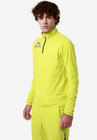 Maglietta atletica a maniche lunghe di un luminoso giallo con colletto alto, zip nera e logo Kappa sul petto. Design in tessuto liscio e elasticizzato.