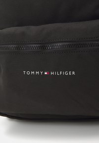 Svart tygryggsäck med en strukturerad yta. Har en dragkedja och "TOMMY HILFIGER"-logotyp i vitt med röda och blå detaljer.