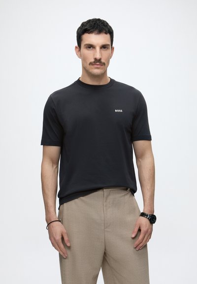 Homme aux cheveux courts et foncés avec une moustache, portant un t-shirt noir "BOSS", un pantalon beige, une montre noire et un bracelet noir, debout devant un fond blanc.
