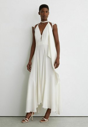 Femme portant une robe blanche fluide sans manches avec un décolleté en V profond et des sandales blanches à talons assorties, debout contre un mur blanc.