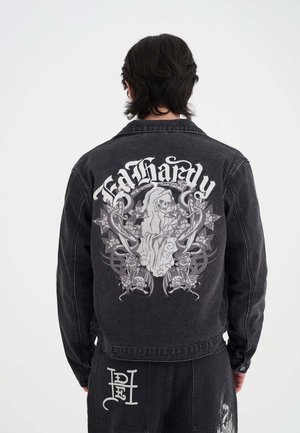 Osoba ubrana w czarną kurtkę dżinsową z detalicznym nadrukiem Kostuchy i napisem "Ed Hardy" na plecach, w połączeniu z czarnymi spodniami z białymi wzorami.