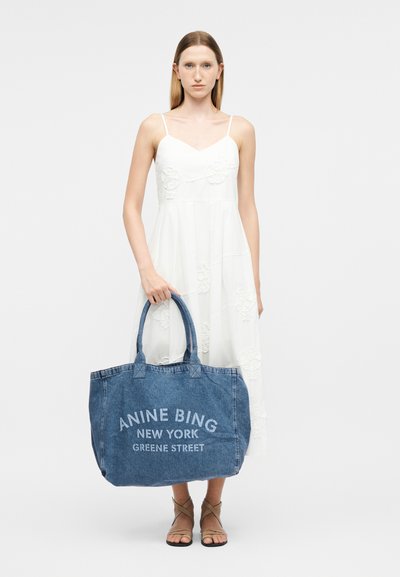 Vrouw in een witte mouwloze jurk met een grote blauwe denim shopper met de tekst "ANINE BING NEW YORK GREENE STREET".