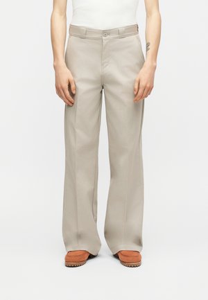 Person wearing beige wide-leg pants and brown shoes, standing against a plain white background, hands relaxed by their sides.