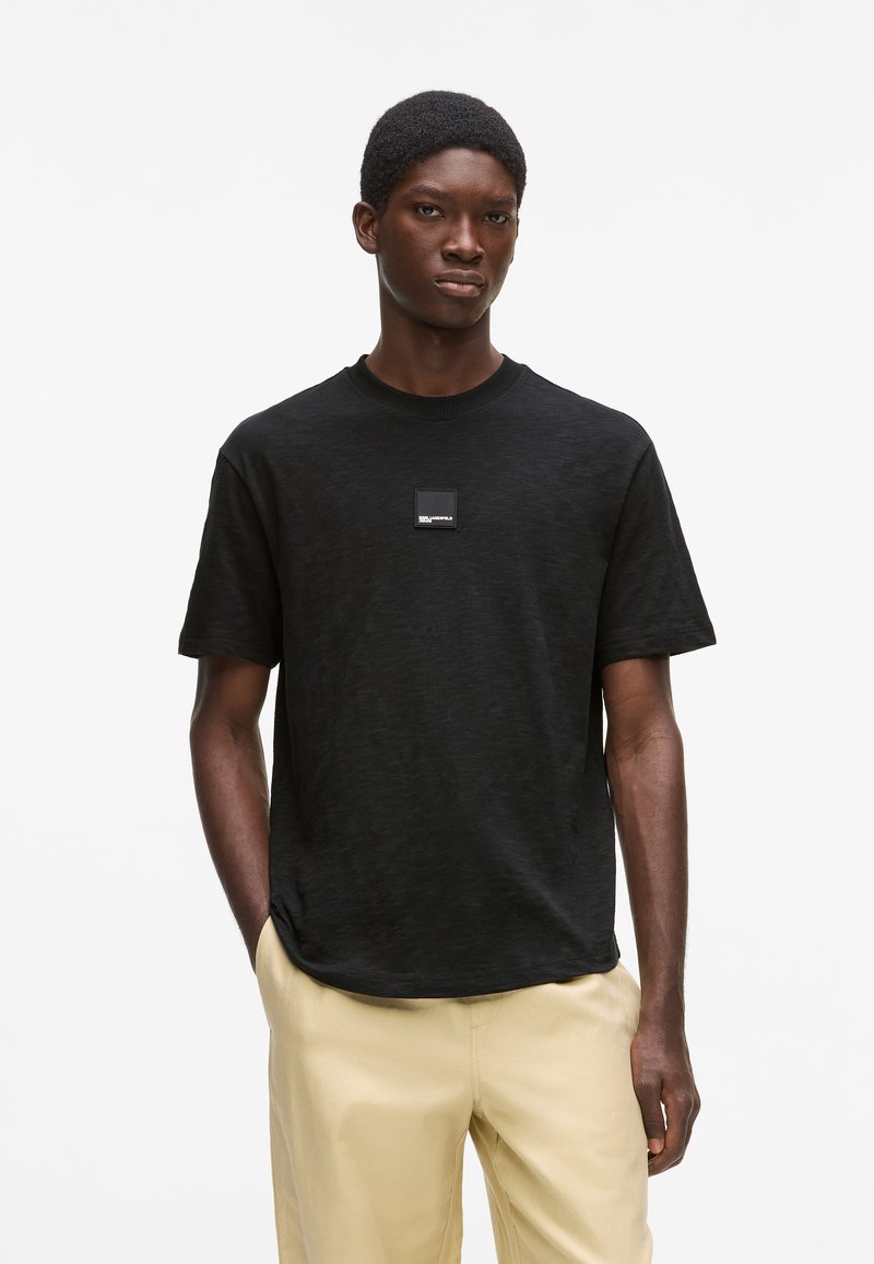 T-shirt noir à col rond avec une texture subtile, manches courtes, et une petite étiquette de marque rectangulaire sur la poitrine, porté avec un pantalon beige.
