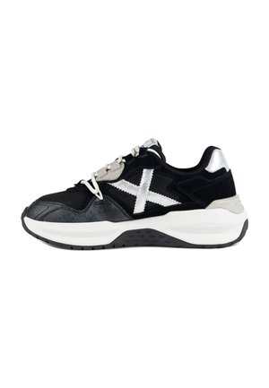 Zapatilla deportiva en blanco y negro con suela blanca gruesa, detalles plateados reflectantes y detalle blanco cruzado en el lateral.