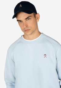 Jasnoniebieska bluza z raglanowymi rękawami i małym bordowym logo. Granatowa czapka baseballowa z logo z przodu. Gładka tekstura.
