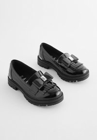 Svarta patentläderloafers med en stor rosettdetalj, fransad front och tjocka gummisulor. Slät textur och glänsande finish.
