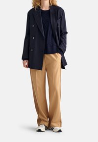 Granatowa marynarka dwurzędowa, teksturowany granatowy sweter z dzianiny, beżowe spodnie o szerokich nogawkach oraz białe trampki z granatowymi akcentami. Gładki, uporządkowany wygląd.