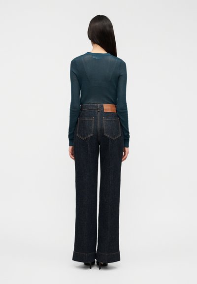 Μακρυμάνικη τοπ σε τιρκουάζ χρώμα και ψηλόμεσο παντελόνι από σκούρο τζιν με φαρδιά μπατζάκια, δερμάτινο ταμπελάκι σε ταμπά απόχρωση, ορατές ραφές και φουσκωτή τελική ραφή.