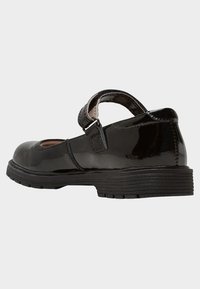 Black patent leather Mary Jane shoe with a Velcro strap and textured rubber sole, shown from the side on a white background.