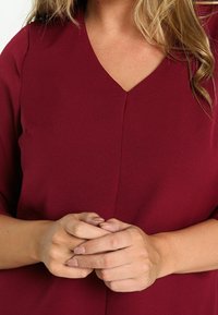 Femme aux cheveux blonds ondulés portant un chemisier bordeaux à col en V, les mains jointes au niveau de la taille.