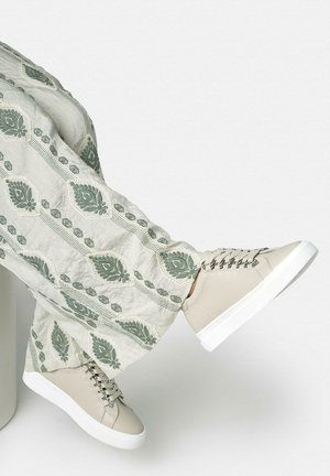 Piernas cruzadas con zapatillas de deporte beige claro con suelas blancas y pantalones anchos con bordados florales verdes sobre tela clara.