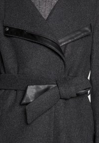 Harmaa villakangastakki, jossa on jäsennelty muotoilu, mustat nahkakoristeet, epäsymmetrinen vetoketju ja vyötäröllä vyö. Teksturoidusta kankaasta ja tyylikkäät viimeistelyt.