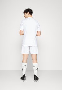 Witte sportshirt en -shorts met zwarte voetbalschoenen en knie hoge witte sokken met zwarte strepen en branding.