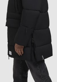 Sort polstret jakke med høj krave, udstyret med lynlåslommer og et logo patch. Stoffet har en glat tekstur og et puffy design.