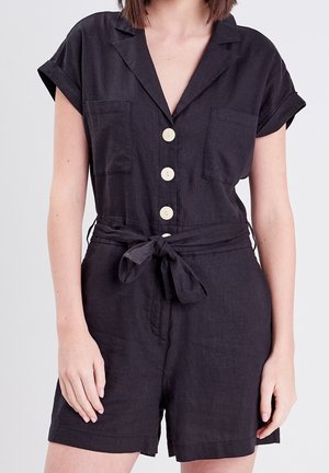 Femme portant une combinaison courte noire à manches courtes avec deux poches poitrine et une ceinture en tissu nouée à la taille.