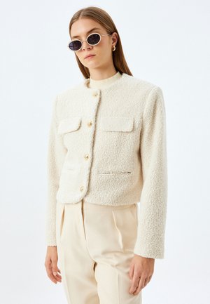 Cropped Jacke aus cremigem Material mit zwei vorderen Taschen und goldenen Knöpfen, kombiniert mit beigefarbenen Hosen und runden, dunklen Sonnenbrillen.