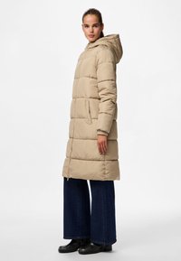 Beige polstret jakke med hette, har glidelås og sidelommer, kombinert med vide mørkeblå jeans og svarte støvler.