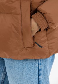 Brun pufferjakke med elastiske manchetter og en sidelomme, fremstillet i et struktureret stof med et subtilt rutenetmønster. Båret over lyseblå jeans.