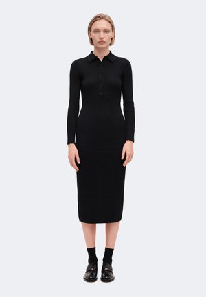Femme aux cheveux blonds courts portant une longue robe noire en maille côtelée avec des boutons, des chaussettes noires et des mocassins noirs, debout devant un fond clair uni.