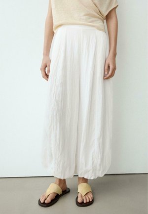 Persona che indossa pantaloni bianchi a vita alta e a gambe larghe e sandali beige, in piedi contro un muro bianco semplice e un pavimento grigio.