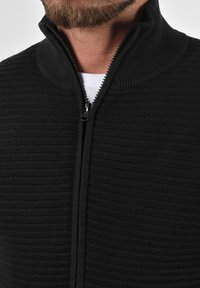 Schwarze Zip-Jacke mit geripptem Struktur, hohem Kragen und metallischem Reißverschluss. Nahaufnahme zeigt die Nähdetails und die Stoffqualität.