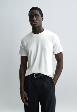 Jauns vīrietis, kurš valkā vienkrāsainu baltu īsroku T-kreklu un melnas bikses ar melnu jostu, stāv ar vienu roku kabatā.