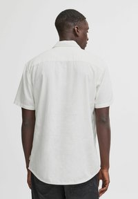 Camisa de manga corta de color beige claro, hecha de una tela suave y texturizada. Presenta un cuello clásico y un dobladillo ligeramente curvado.