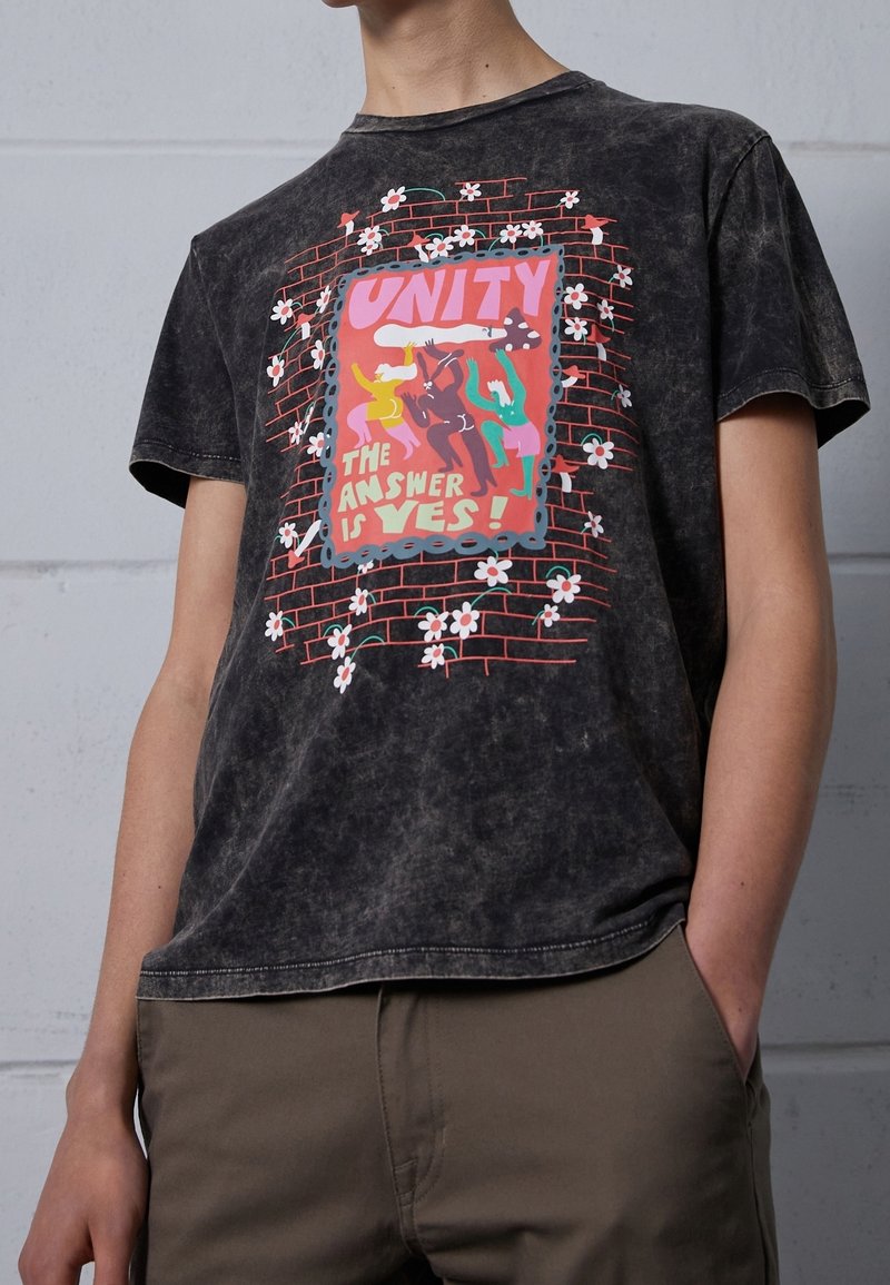 Personne portant un t-shirt noir délavé avec un graphique coloré représentant des figures dansantes, des fleurs, un mur de briques, et le texte "L'UNITÉ LA RÉPONSE EST OUI !"