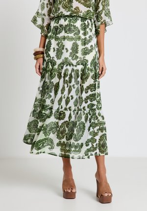 Jupe blanche fluide avec imprimé cachemire vert, associée à des sandales plateformes marron épaisses et des bracelets empilés au poignet gauche.