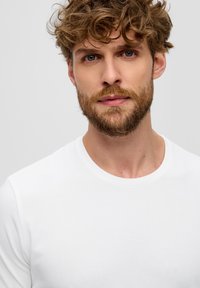 Camisa blanca de manga corta con escote redondo, textura suave y corte casual, llevada por un hombre con cabello castaño claro desordenado y barba.