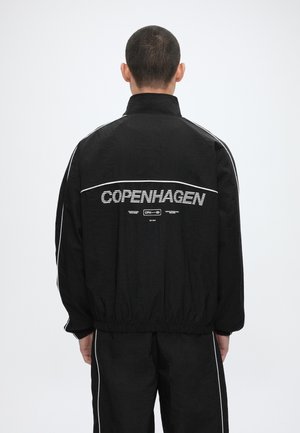 Muž v čiernej bunde a nohaviciach s bielym lemovaním, na chrbte nápis „Copenhagen“, otočený chrbtom k jednoliatemu svetlému pozadiu.