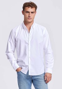 Man in een wit overhemden en lichtblauwe jeans, staande met één hand in zijn zak tegen een effen achtergrond.