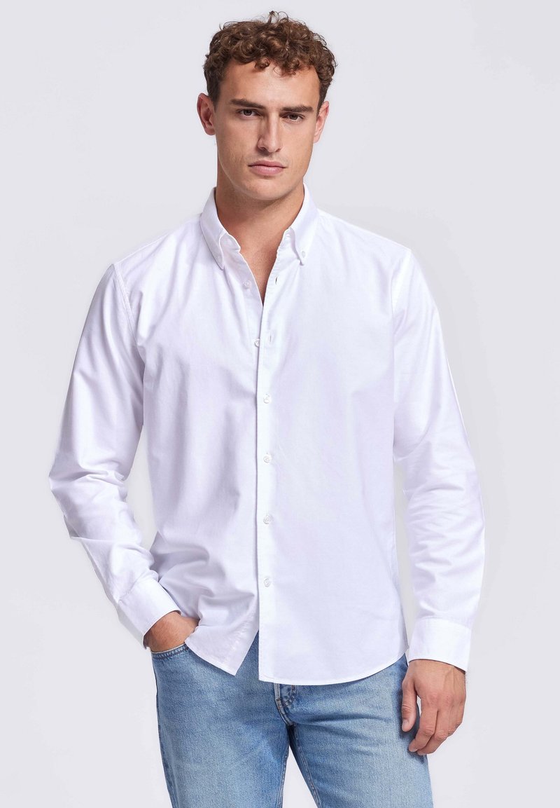 Man in een wit overhemden en lichtblauwe jeans, staande met één hand in zijn zak tegen een effen achtergrond.