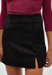 Mini falda de ante negra con una abertura lateral y seis botones decorativos a lo largo de la costura izquierda; textura suave y diseño ajustado.