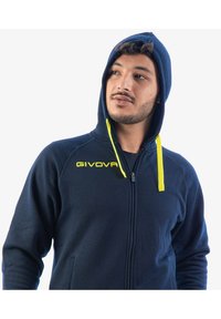 Uomo che indossa una felpa con zip blu navy con coulisse gialla e logo "GIVOVA", che guarda leggermente di lato su uno sfondo bianco.
