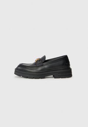 Mocassin en cuir noir avec une semelle en caoutchouc épaisse, présentant un accent doré sur la sangle et une finition lisse.