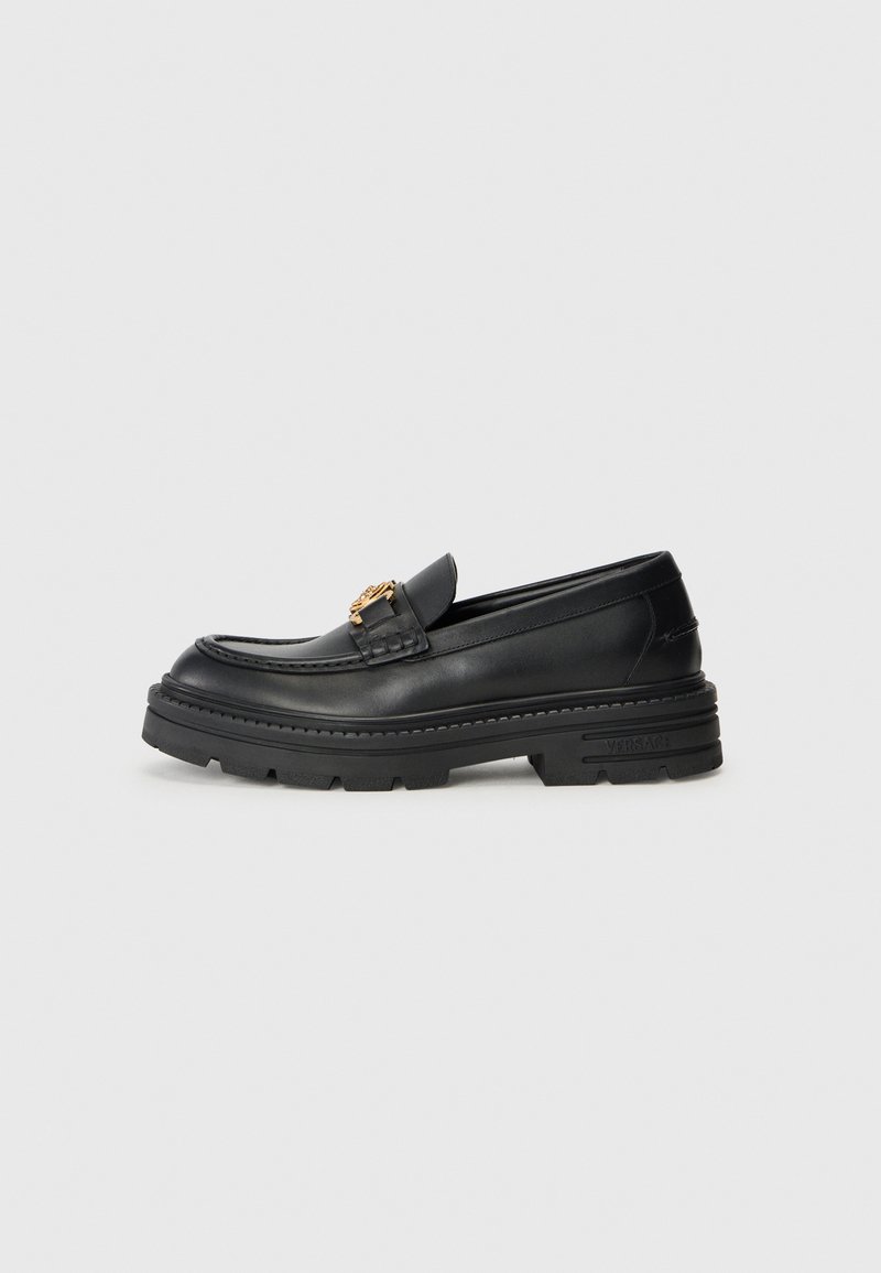 Mocassin en cuir noir avec une semelle en caoutchouc épaisse, présentant un accent doré sur la sangle et une finition lisse.