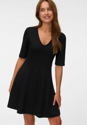 Femme aux longs cheveux bruns souriante, portant une robe noire ajustée avec des manches aux coudes et un décolleté en V, debout devant un fond uni.